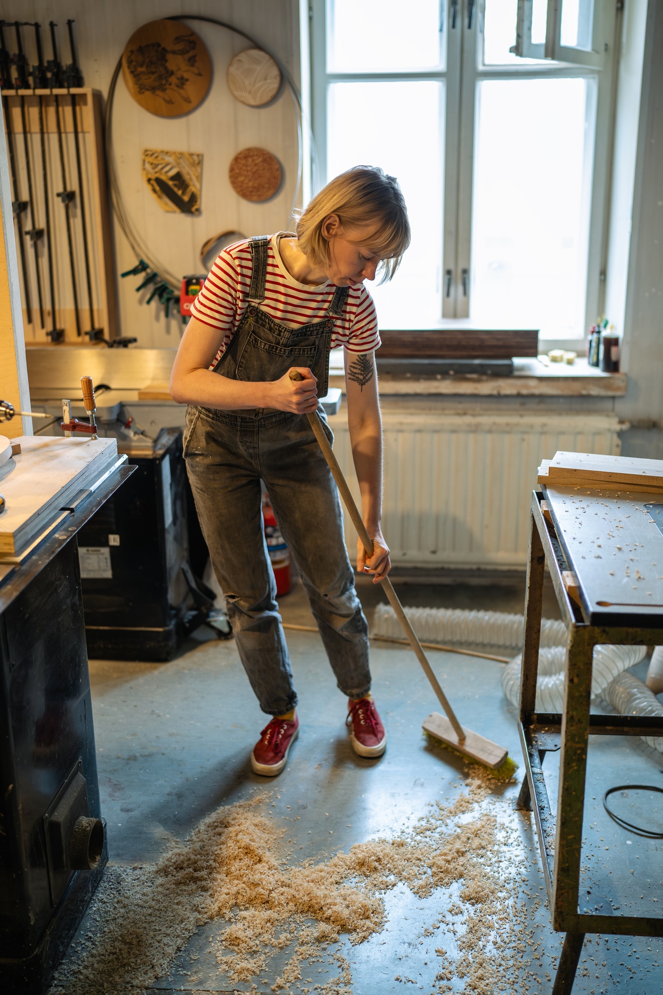 Carpenter woman cleaning up workshop after work. Handywoman sweep sawdust and ventilating for health