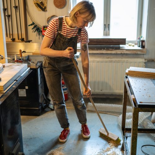 Carpenter woman cleaning up workshop after work. Handywoman sweep sawdust and ventilating for health
